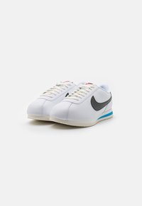Fehér bőr sportcipők fekete swoosh-sal, kék akcentussal a sarkon és texturált gumitalppal. A fűző bézs színű.