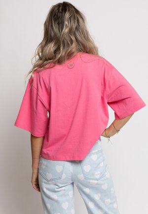 Vrouw met lang golvend haar draagt een loszittend roze T-shirt en lichtblauwe spijkerbroek met witte hartjespatronen, kijkt weg en heeft één hand op haar heup.