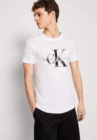 Balta kokvilnas t-kreklā ir drosmīgs melns "CK" logo un teksts "Calvin Klein Jeans", ar apaļu kakla izgriezumu un īsām piedurknēm. Gluda tekstūra.