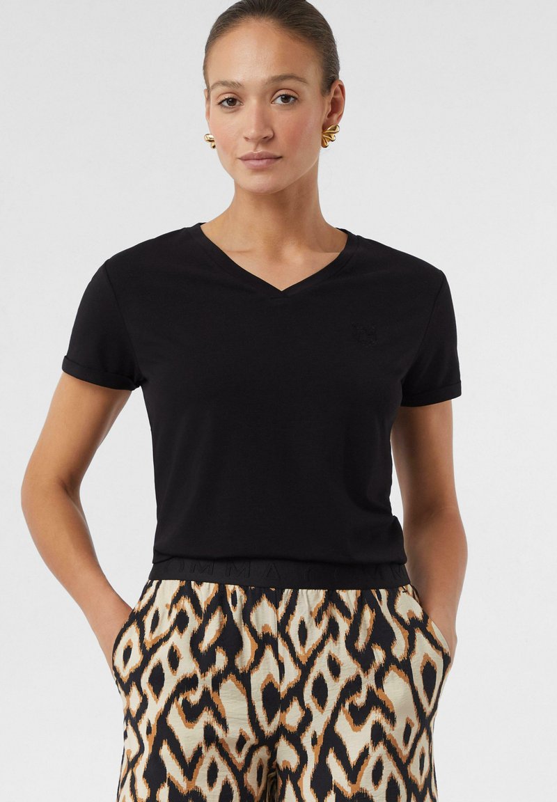 Zwarte T-shirt met korte mouwen en V-hals, gemaakt van zachte stof, gedragen met beige en zwarte pantalon met een patroon dat een abstracte, dierlijke uitstraling heeft.