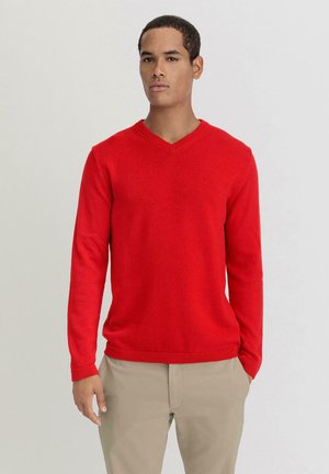 Roter V-Ausschnitt-Pullover mit langen Ärmeln, gerippten Bündchen und Saum, aus einem weichen Stoff gefertigt. Kombiniert mit beigen Hosen.
