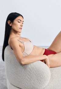 Lichtbeige beha met een lichte padding en dunne bandjes, gecombineerd met een dieprode bikini onderkant. Model ligt achterover op een textuurrijke, ronde beige stoel.