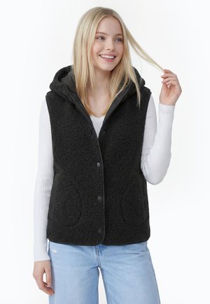 Junge Frau lächelt, trägt eine schwarze Fleece-ärmellose Jacke mit Kapuze über einem weißen, gerippten Langarmshirt und hellblauen Jeans.