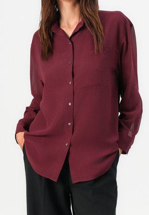 Woman wearing a long-sleeve, buttoned burgundy blouse with a collar and black pants, standing hands in pockets against a white background.