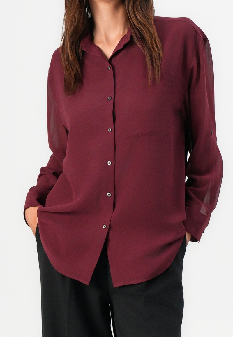 Femme portant un chemisier bordeaux à manches longues, boutonné, avec un col et un pantalon noir, debout les mains dans les poches sur fond blanc.