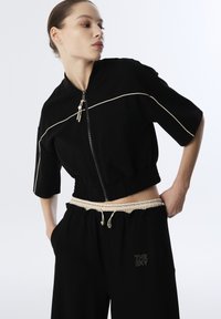 Black cropped jacket with short sleeves, elastic waistband, and cream trim. Paired with black pants featuring a light drawstring and logo detail.