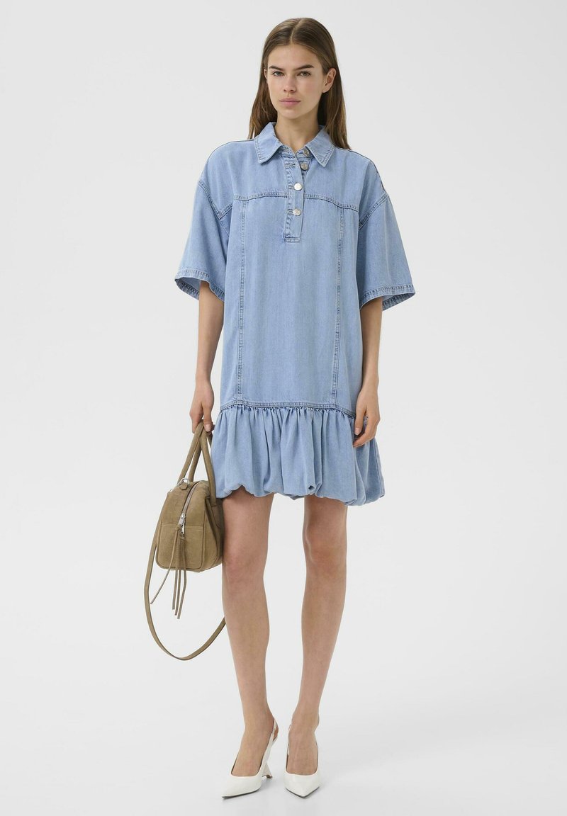 Vrouw draagt een lichtblauwe denim jurk met korte mouwen en een geplooide zoom, witte hakken, houdt een beige handtas vast en staat tegen een effen witte achtergrond.