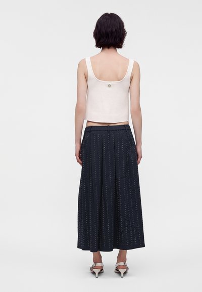 Γυναίκα που φοράει λευκό αμάνικο crop top, σκούρο μπλε παντελόνι με φαρδιά πόδια και μικρές βούλες, και λευκά ψηλοτάκουνα σανδάλια ανοιχτά στη μύτη, απεικονιζόμενη από πίσω.