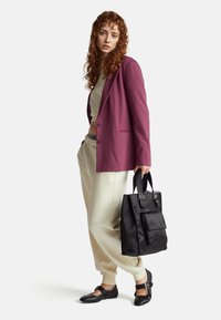 Borsa a tracolla nera con manici, blazer viola, top crema e pantaloni jogger chiari. Modella in piedi con capelli ricci, sfondo neutro.