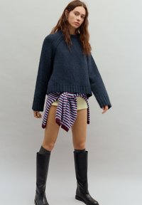 Tengerészkék kötött pulóver bordázott mandzsettákkal, világos színű rövidnadrággal párosítva, csíkos bordó és krém színű kendővel, térdig érő fekete csizmával.