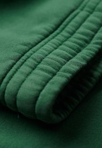 Zielony materiał o miękkiej teksturze, z pikowanym wzorem wzdłuż złożonej krawędzi, prezentujący wiele warstw i gładkie wykończenie.