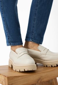 Dikke witte leren loafers met een gestructureerde afwerking en beige rubberen zolen, gecombineerd met blauwe spijkerbroeken, rustend op een houten oppervlak.