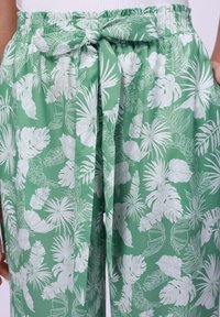 Pantalones de tela ligera verde con un patrón de hojas blancas tropicales, cinturilla elástica con un detalle de lazo, y bolsillos laterales.