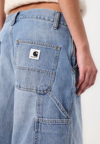 Ljusblå denimbyxor har en stor bakficka med en vit logotyp och synliga sömmar. Tyget har en något sliten struktur.