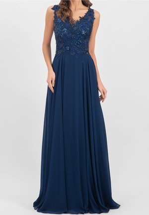 Kobieta ubrana w granatową wieczorową suknię bez rękawów z kwiecistym koronkowym i cekinowym górnym elementem oraz zwiewną spódnicą z szyfonu, stojąca na jednolitym tle.