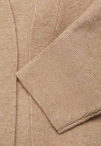 Manica e sezione del corpo di un morbido pullover lavorato a maglia color beige, con dettagli a coste e cuciture.