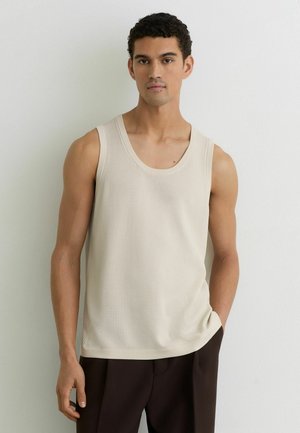 Noor mees lühikeste lokkis juustega, kellel on seljas varrukateta beež kudumist kampsun ja tume püksid, seisab ühevärvse valge seina ees.