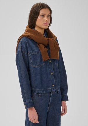 Jeune femme portant une veste en jean bleue et un jean avec un pull marron drapé sur ses épaules, sur un fond gris uni.