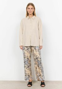 Beige Hemd mit Knöpfen und einer einzelnen Brusttasche, kombiniert mit lockeren, gemusterten Hosen in Beige, Grau und Schwarz. Schwarze Sandalen mit offenen Zehen.