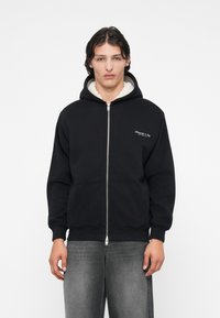 Sweat à zip noir avec une doublure en polaire blanche et un logo brodé sur la poitrine gauche, associé à un jean gris et un fond uni.