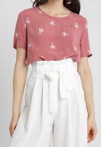 Kort pink top med korte ærmer og hvidt blomstermønster, parret med højtaljede hvide bukser med en bundet sløjfedetalje.