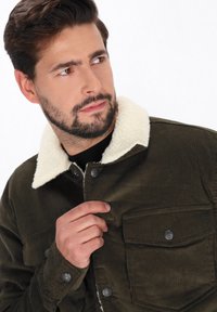 Jachetă din corderoy verde închis, cu guler din blană sintetică crem, având închidere cu nasturi și două buzunare la piept. Detaliu de țesătură vizibil.