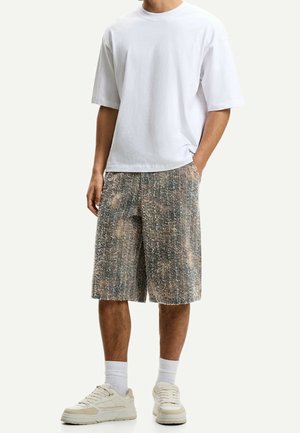 Man die een effen wit oversized t-shirt draagt, gestructureerde meerkleurige knielange korte broek, witte sokken en beige sneakers, staand met één hand in de zak.