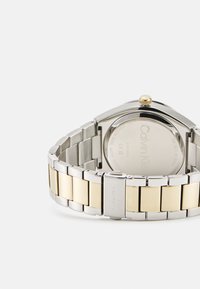 Reloj de acero inoxidable y metal en tono dorado con caja rectangular, eslabones de pulsera plateados y corona dorada. Logotipo de la marca grabado en el cierre.