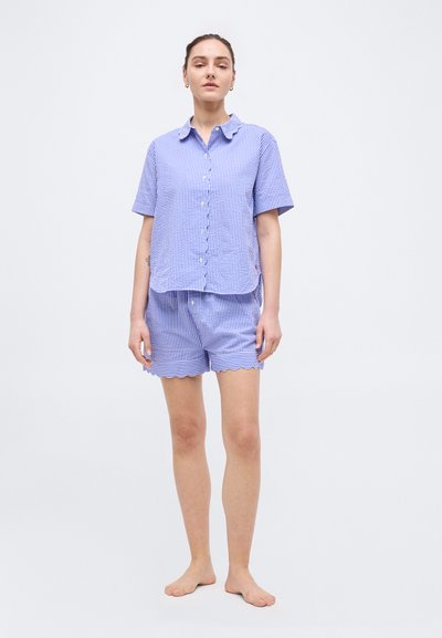 Γυναίκα που στέκεται ξυπόλυτη, φορώντας μπλε και λευκό ριγέ πουκάμισο με κοντά μανίκια και κουμπιά και ταιριαστό σορτς με κυματιστό τελείωμα.