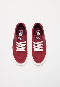 Røde ruskindsneakers med hvide snørebånd, perforeret tåhætte, polstret krave og kontrastfuld grå side-stribe. Vans-logoet er synligt på tungen.