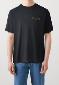 Mężczyzna noszący czarną koszulkę z krótkim rękawem Abercrombie & Fitch z małym złotym logo na lewej piersi oraz niebieskie dżinsy.