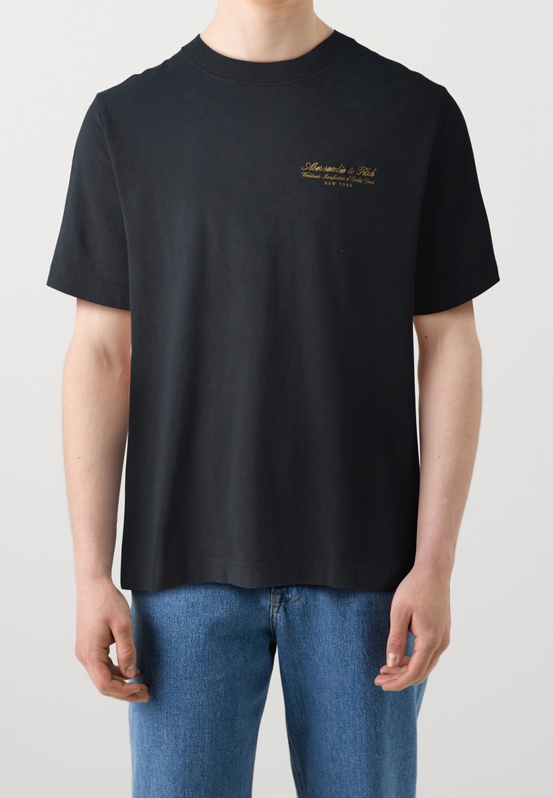 Mężczyzna noszący czarną koszulkę z krótkim rękawem Abercrombie & Fitch z małym złotym logo na lewej piersi oraz niebieskie dżinsy.