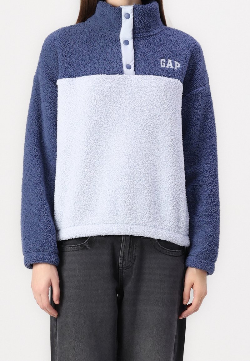 Personne portant un pull polaire bleu et blanc avec boutons-pression et logo GAP, associé à un jean noir, debout devant un fond uni.