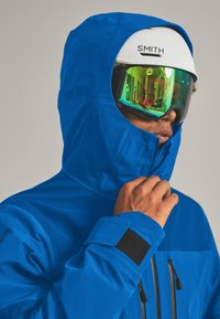 Blaue wasserdichte Jacke mit Kapuze, schwarzen Klettverschlussakzenten, zipfbaren Taschen und einem strukturierten Stoff. Ausgestattet mit einem weißen Helm und grüntönenden Schutzbrille.