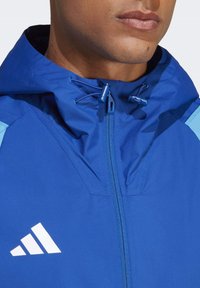 Blaue Kapuzenjacke mit Reißverschluss, auffälligen hellblauen Akzenten und einem weißen Logo auf der linken Seite. Texturiertstoff und verstellbare Kapuze.