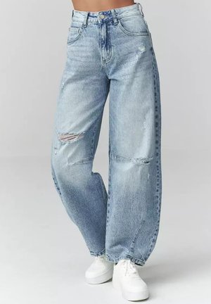 Jasnoniebieskie jeansy z wysokim stanem i szerokimi nogawkami z przetarciami na kolanach, noszone wraz z białymi trampkami na platformie, pokazane od talii do stóp na jednolitym tle.