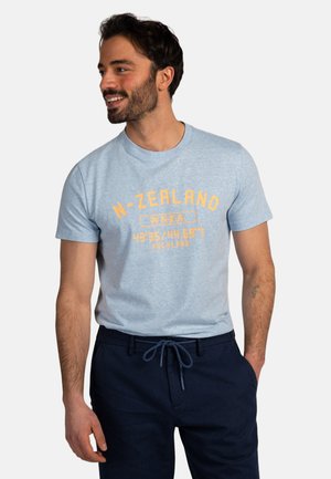 Lachende man met donker haar en baard draagt een lichtblauw T-shirt met de tekst "N-ZEALAND" en donkerblauwe broek met handen in de zakken.