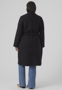 Svart ullblande kåpe med belte, lange ermer og spiss krage. Bært over blå vide jeans med et minimalistisk design.