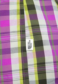 Pestrý kostkovaný materiál se fialovými, zelenými a šedými pruhy. Obsahuje bílou štítku s logem "The North Face".