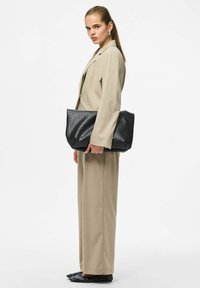 Melnā izšūtā somiņa ar teksturētu apdari, ko tur modeļa rokās. Viņa valkā bēša krāsas kostīmu ar piegrieztu siluetu un gludām biksēm.