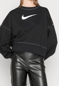 Sudadera negra recortada con logo blanco de Nike, con mangas largas, cuello redondo y detalles de costura en contraste. Combinada con pantalones de cuero negros.