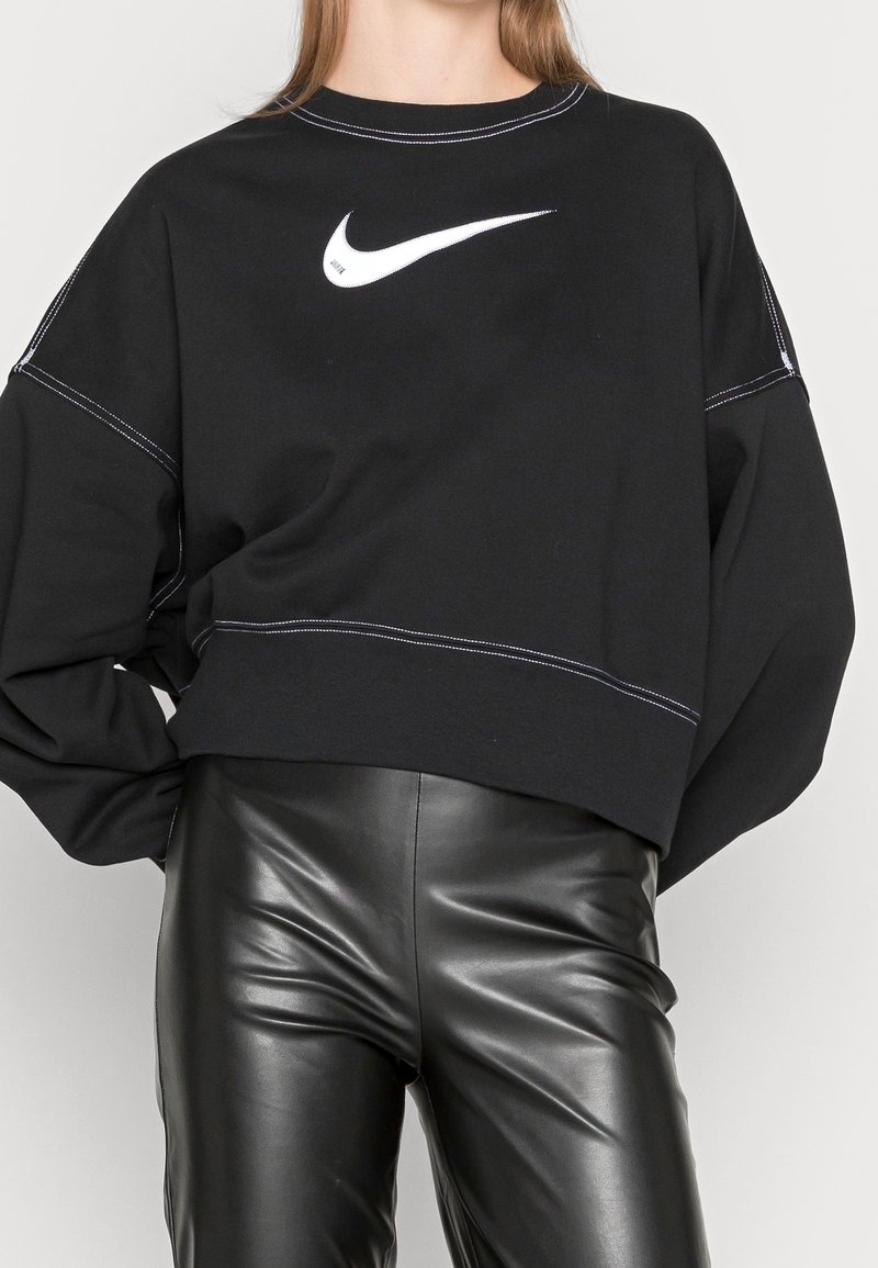 Sudadera negra recortada con logo blanco de Nike, con mangas largas, cuello redondo y detalles de costura en contraste. Combinada con pantalones de cuero negros.