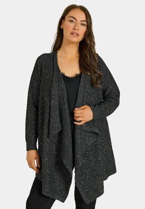 Femme portant un cardigan noir scintillant ouvert sur un haut noir, debout, une main tenant le cardigan près de la poitrine.