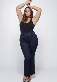 Zwart mouwloos bodysuit en donkerblauwe flared jeans met zichtbare stiksels. Model staat geposeerd met opgetilde armen, waardoor de outfit goed zichtbaar is.