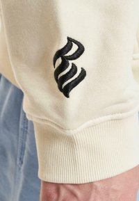 Manica di felpa color crema con polsini a coste e design del logo ricamato in nero. Texture morbida e vestibilità leggermente rilassata.