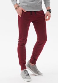 Bordeaux joggers met een taps toelopende pasvorm hebben zijzakken, elastische boorden en een gladde textuur, gecombineerd met lichtgrijze sneakers.