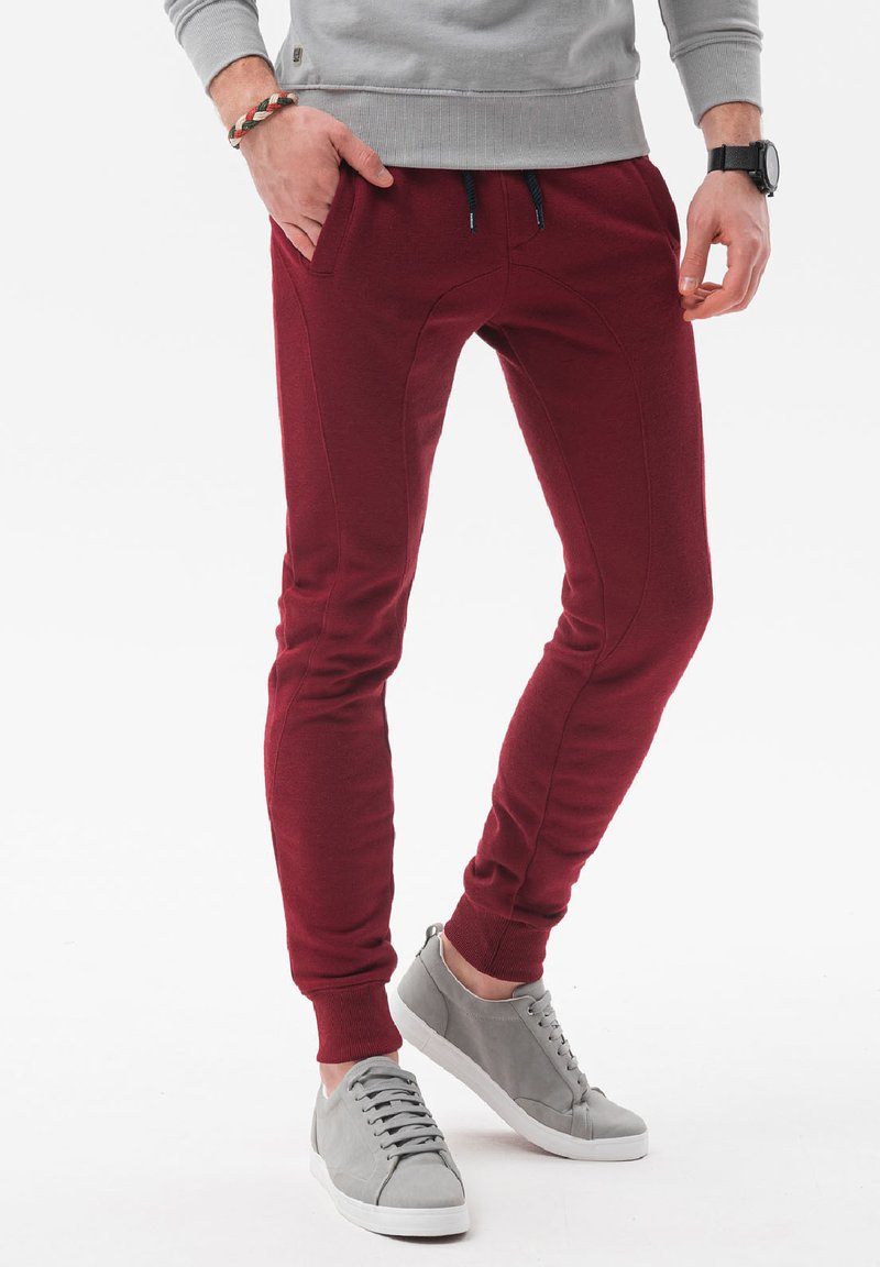 Bordeaux joggers met een taps toelopende pasvorm hebben zijzakken, elastische boorden en een gladde textuur, gecombineerd met lichtgrijze sneakers.