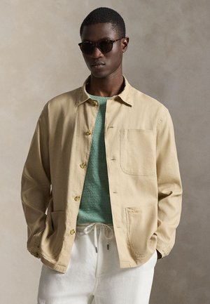 Άνδρας που φοράει σκουρόχρωμα γυαλιά ηλίου, μπεζ σακάκι με κουμπιά, πράσινο πλεκτό πουλόβερ και λευκό παντελόνι με κορδόνι, σε ουδέτερο φόντο.