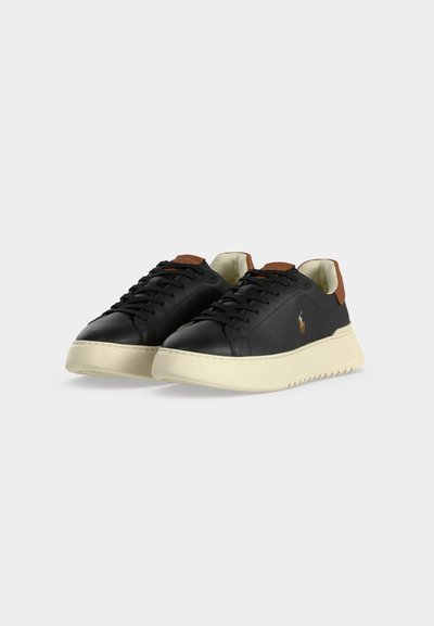 Baskets basses en cuir noir avec semelles épaisses couleur crème, languettes marron au talon, lacets noirs et petit logo doré sur le côté.