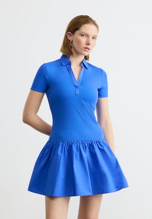 Kvinde iført en tætsiddende blå polokjole med krave og korte ærmer, stående mod en ensfarvet hvid baggrund.